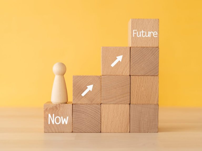 wooden figure on a wooden block that says "Now" with more wooden blocks arranged as steps up to a wooden block that says "Future"